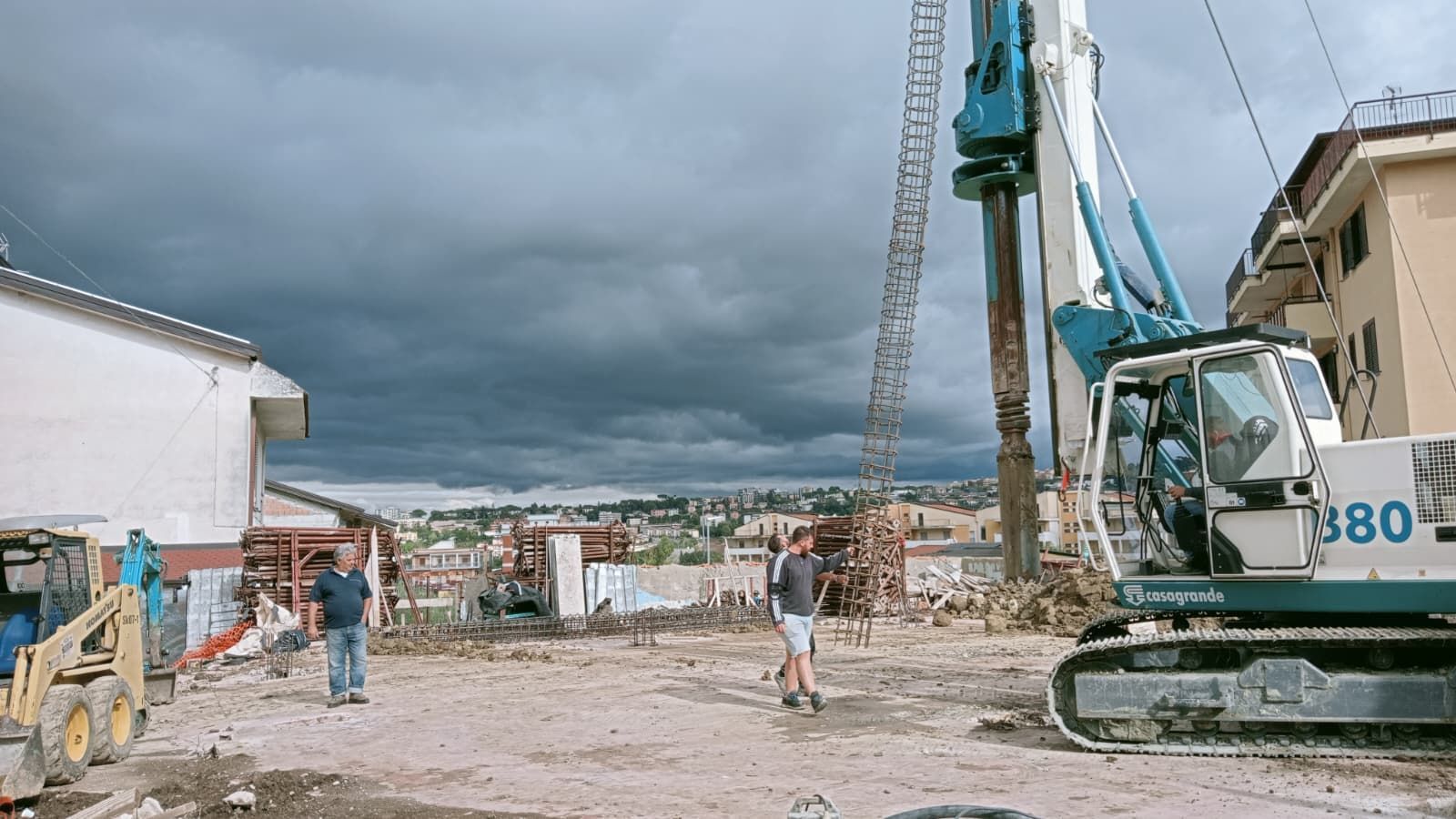 Cantiere edile con operai e una gru accanto a edifici danneggiati sotto un cielo nuvoloso.