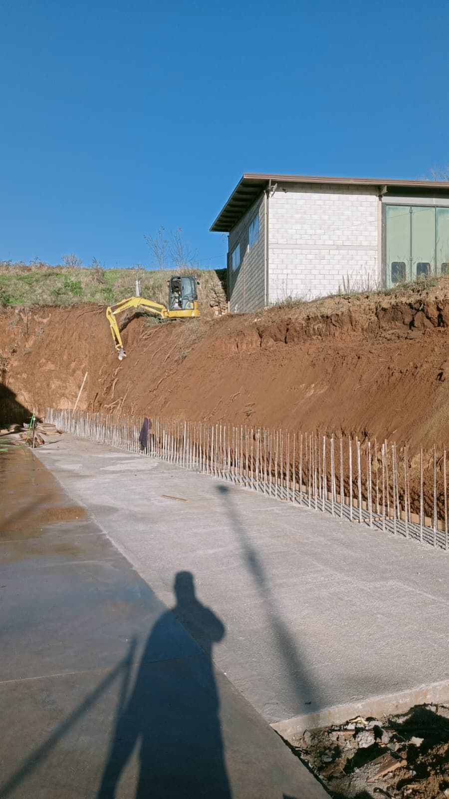 Cantiere edile con escavatore che livella un pendio di terra accanto a una strada asfaltata e un piccolo edificio sotto un cielo azzurro e limpido.