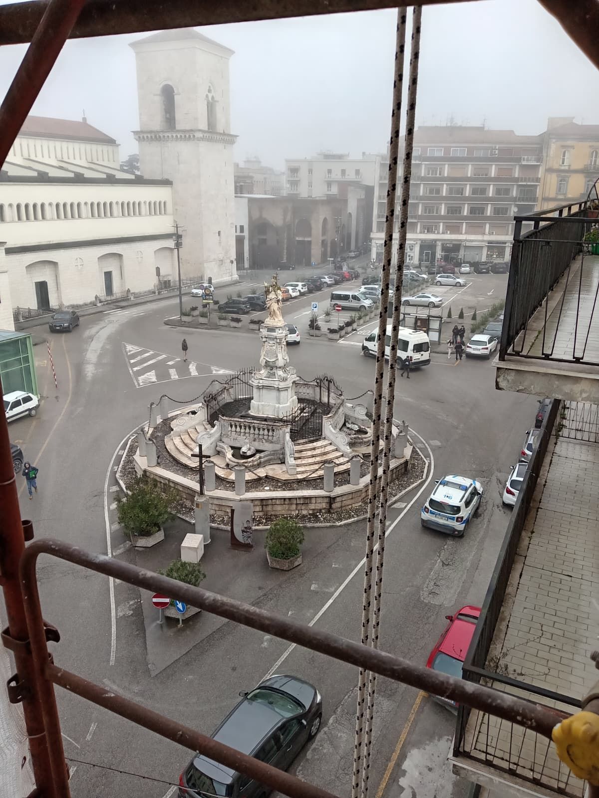 Vista nebbiosa di una piazza cittadina con fontana centrale, edifici circostanti e auto parcheggiate, ripresa da un balcone sopraelevato.