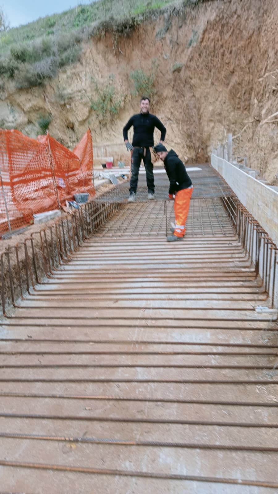Due operai su una passerella di legno inclinata, uno dei quali si sporge in avanti vicino a una rete di sicurezza arancione su un pendio sterrato.