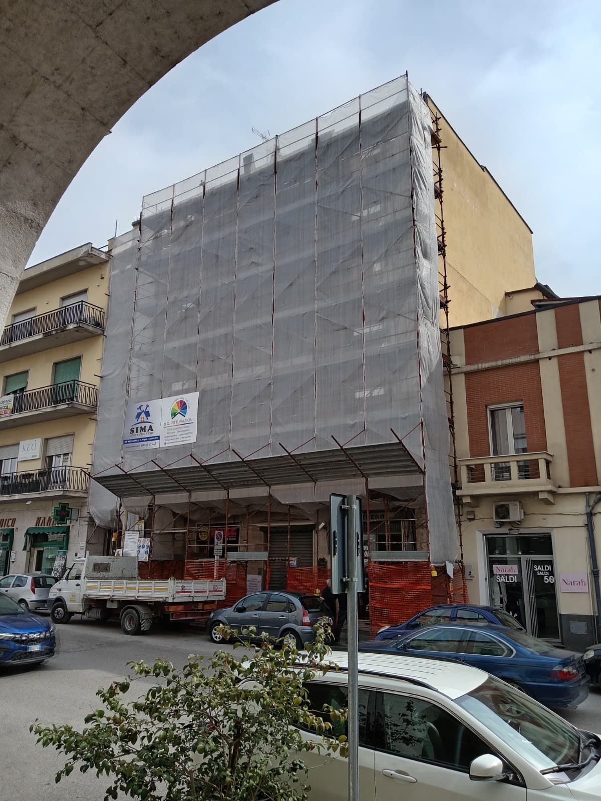 Un edificio con impalcature e copertura in rete grigia si erge su una strada cittadina, con auto parcheggiate al di sotto.