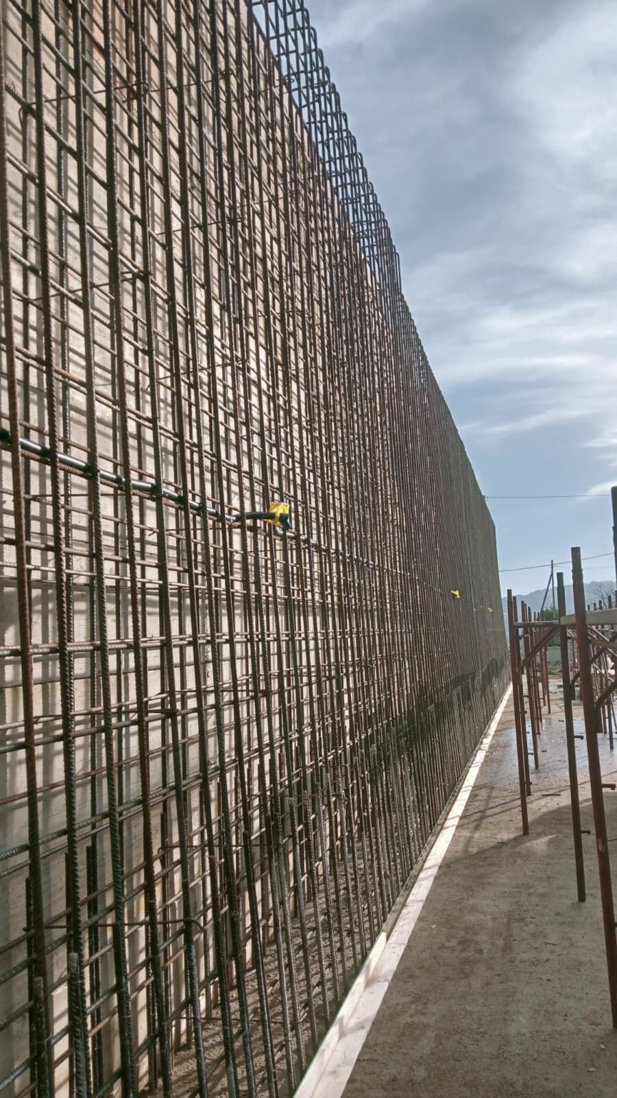 Un lungo muro di barre d'armatura lungo una passerella di cemento in un cantiere edile sotto un cielo nuvoloso.