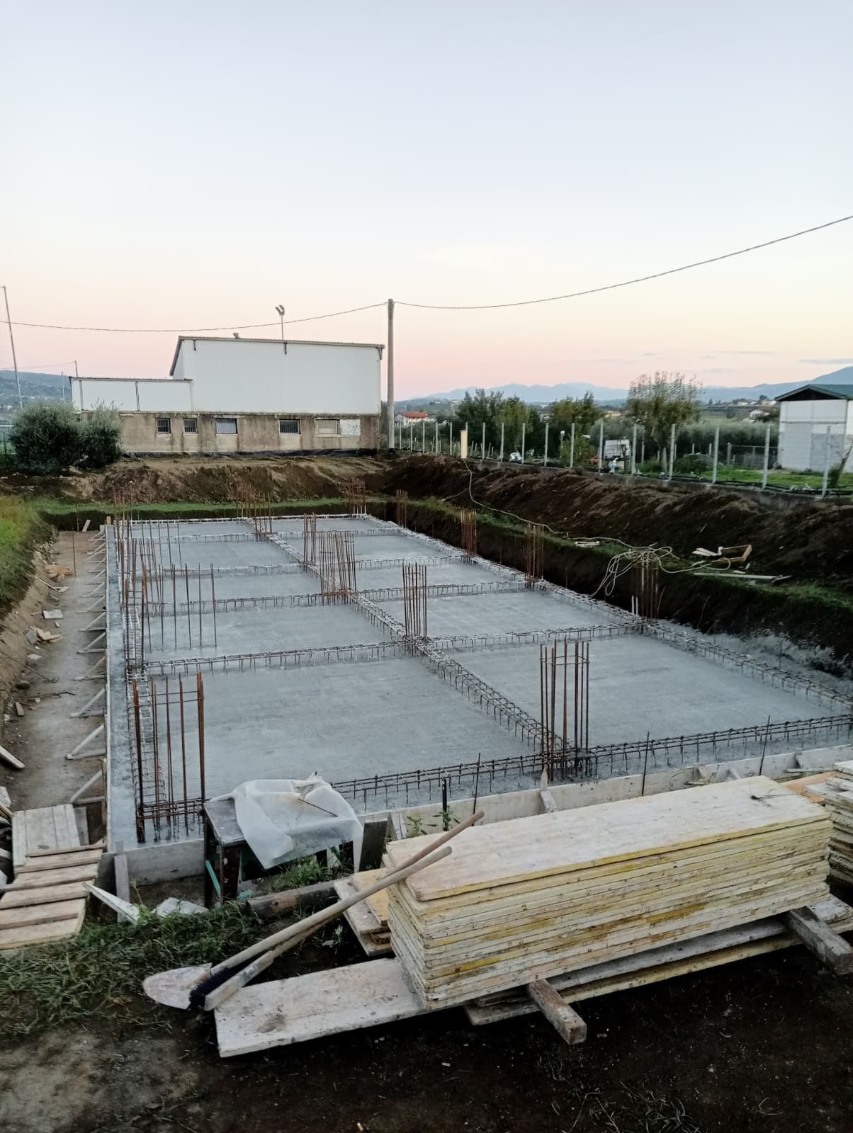 Fondamenta in cemento in costruzione al crepuscolo, con barre d'armatura, legname e case vicine.