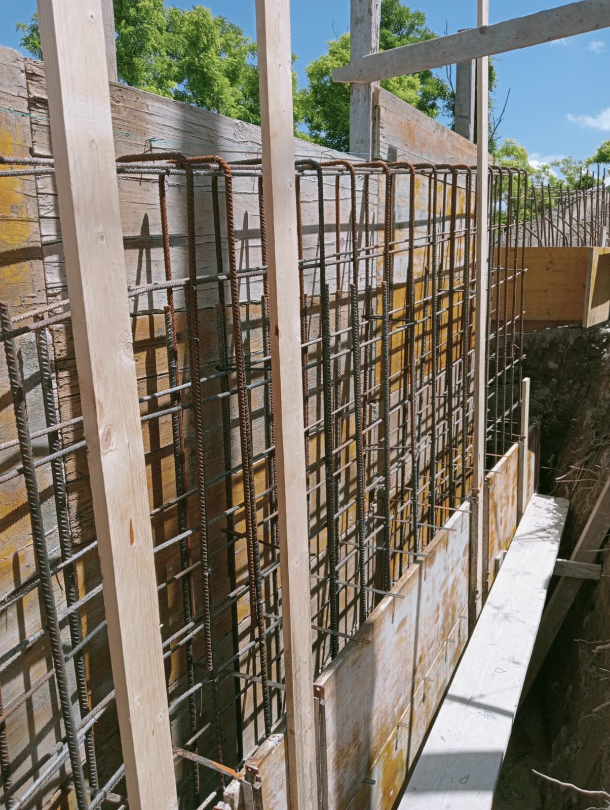 Muro di cemento parzialmente costruito con armature a vista accanto a uno stretto passaggio pedonale sotto un cielo azzurro.