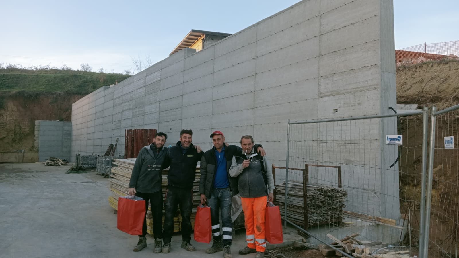 Gruppo di operai con giacche scure e pantaloni arancioni in piedi accanto a un muro di cemento in un cantiere.