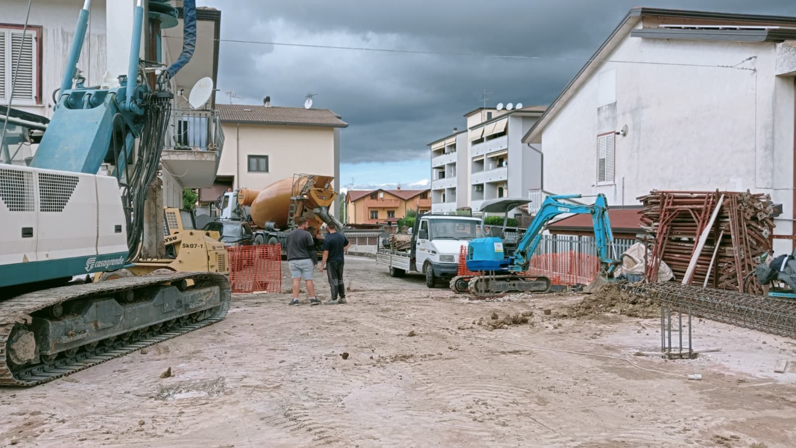 Cantiere edile con escavatore, operai e materiali sparsi su una strada fangosa sotto un cielo nuvoloso.