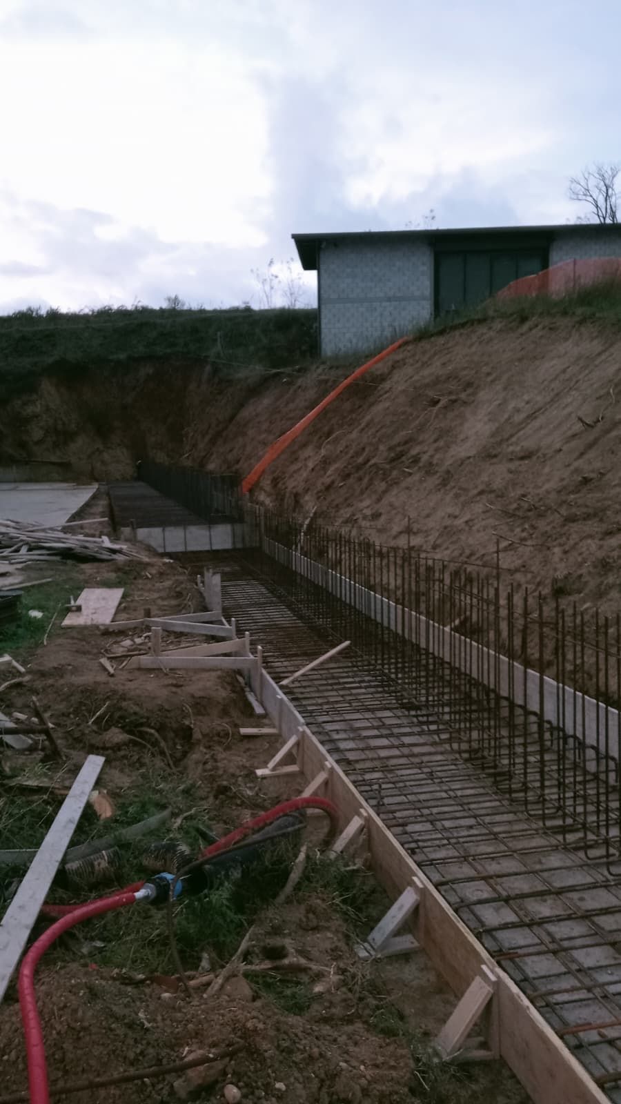 Scavo ripido in un cantiere edile con armature in ferro a vista, casseforme in legno e un piccolo edificio in cima al pendio.