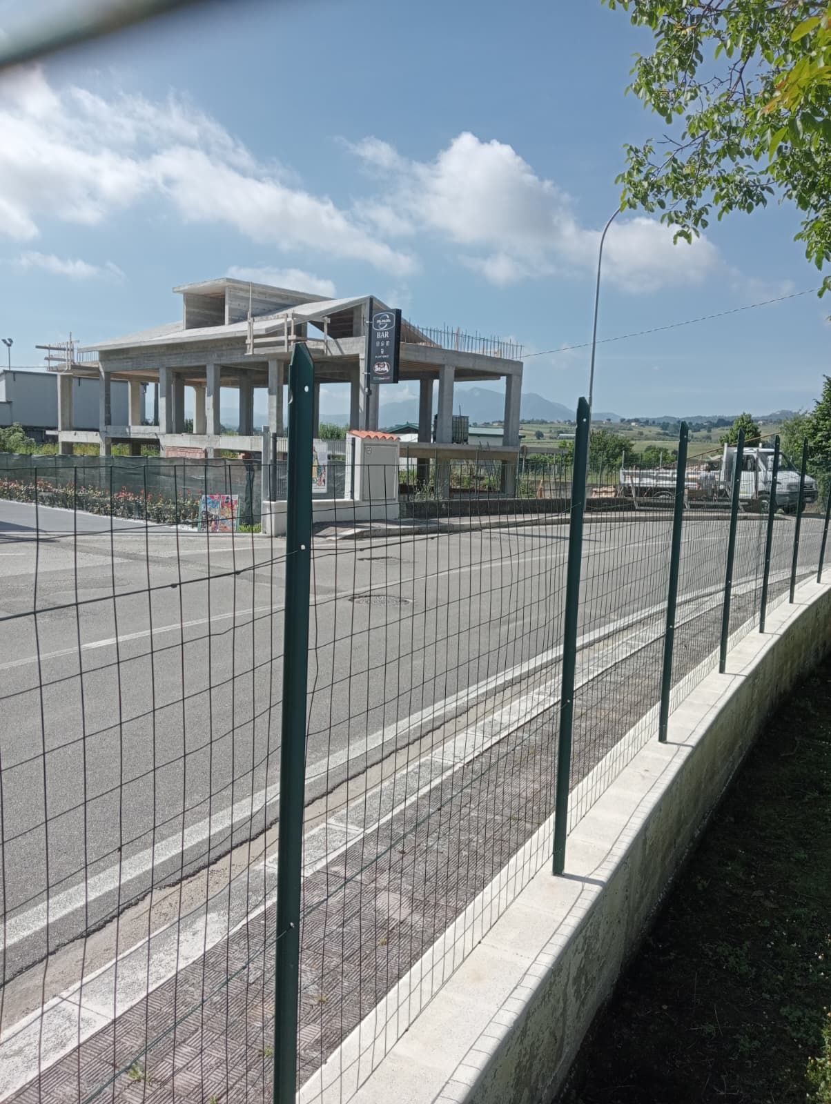 Terreno pavimentato vuoto accanto a un edificio in cemento non finito e recintato, sotto un cielo azzurro.