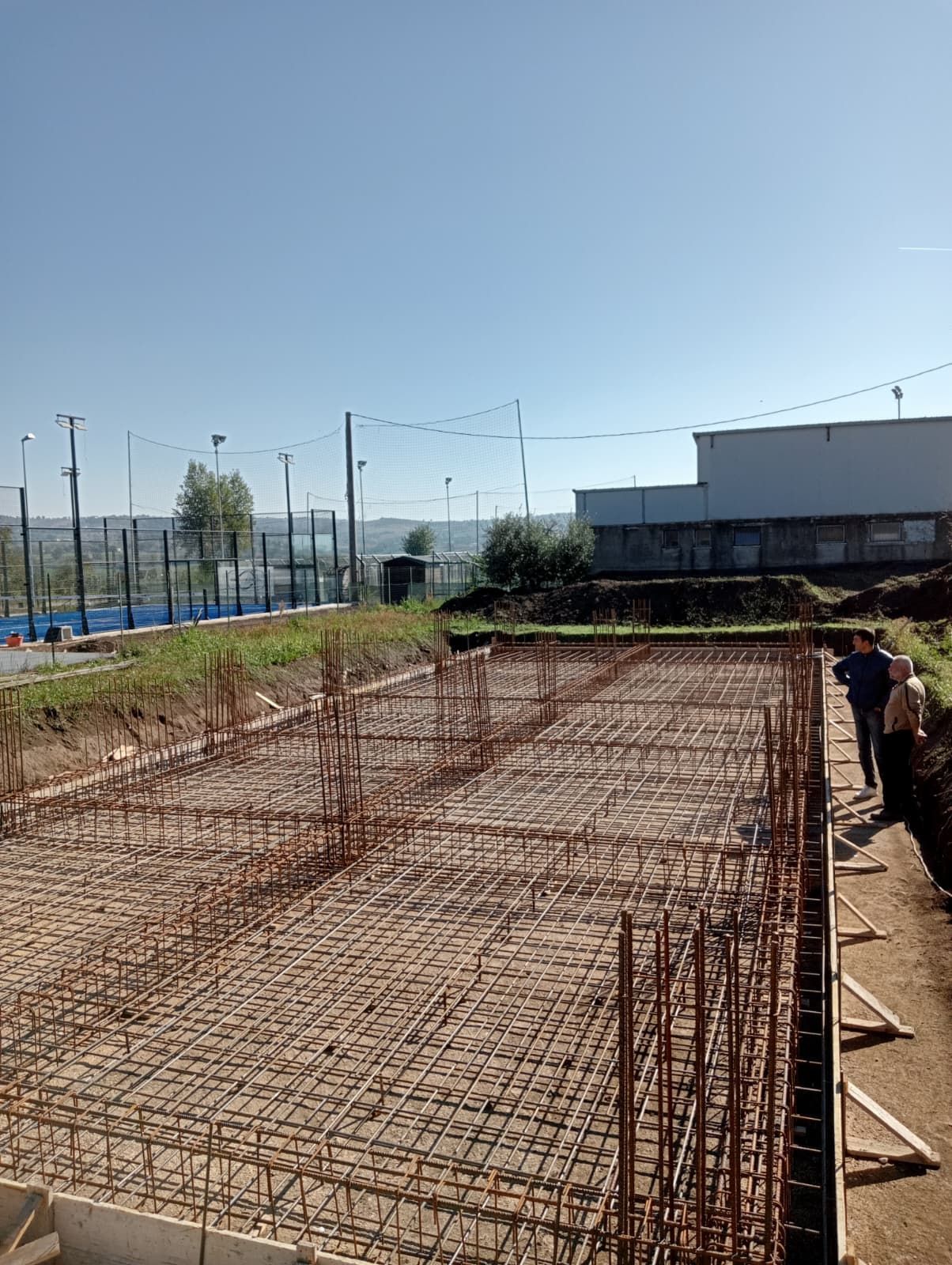 Cantiere edile con griglia di fondazione in tondini d'acciaio e operai lungo il bordo destro sotto un cielo sereno.