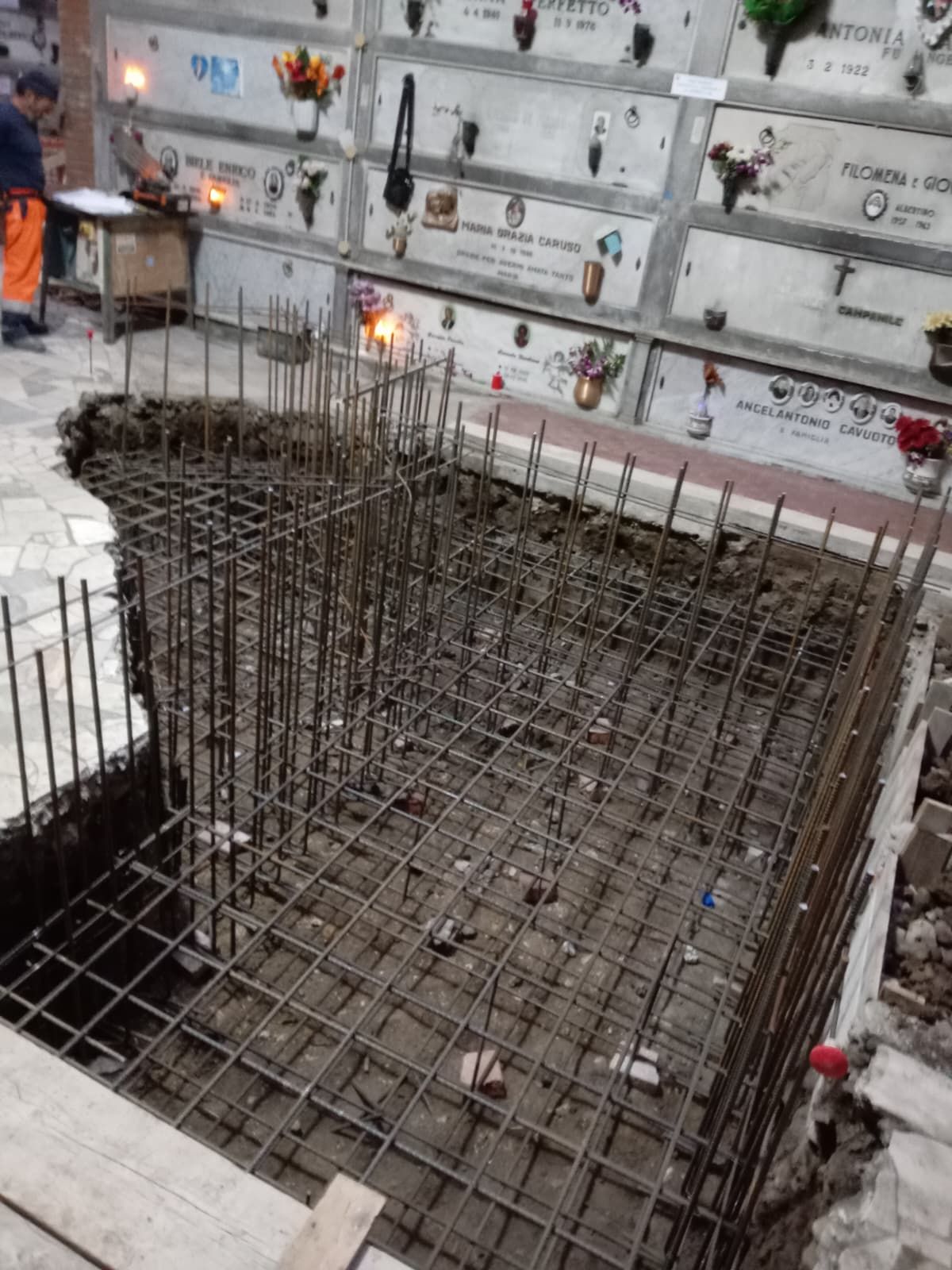 Gabbia di fondazione in tondini di ferro in una fossa di cantiere, con pareti piastrellate di bianco e piccole candele nelle vicinanze.