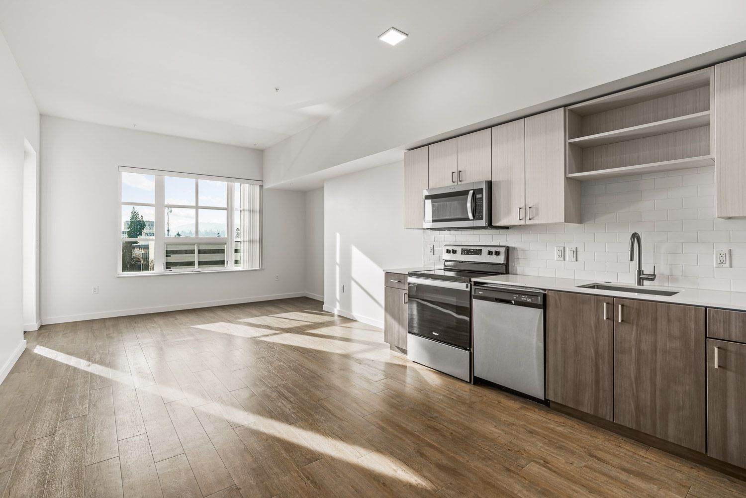 Spacious, light-filled apartment at Gordon Lofts in Eugene, OR, featuring a modern kitchen with stainless steel appliances, two-tone cabinetry, open shelving, and a white subway tile backsplash. Warm wood-style flooring extends through the open living area, which is illuminated by a large window offering views of the city and hills beyond.