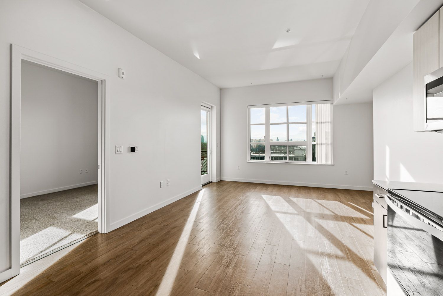 Bright and modern 1-bedroom apartment at Gordon Lofts in the 5th Street Public Market, Eugene, OR. The unit features large windows with natural light, wood-style flooring in the main living area, a carpeted bedroom and a sleek kitchen with stainless steel appliances.