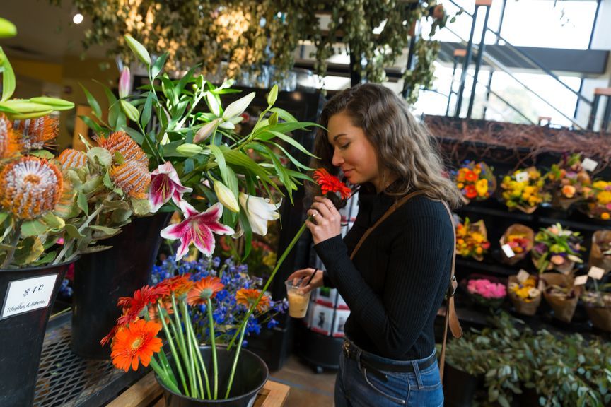 A woman holding an iced coffee and smelling a vibrant red flower inside Rhythm & Blooms at 5th Street Public Market. She is surrounded by colorful flower arrangements, including lilies, proteas, and gerberas, displayed in buckets.
