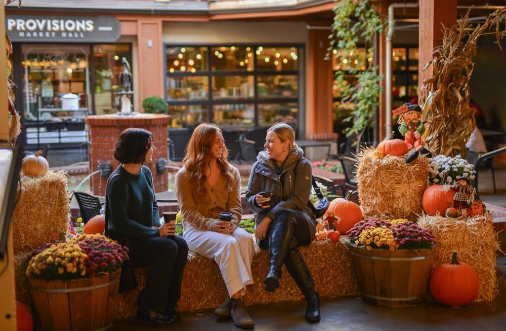 Three women sitting on straw bales decorated with pumpkins, flowers, and fall-themed ornaments in the Market Core of 5th Street Public Market. They are enjoying drinks and conversation in a cozy, festive autumn setting with Provisions Market Hall visible in the background.