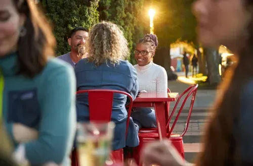 Group of people enjoying an outdoor dining experience at 5th Street Public Market, seated at a vibrant red table surrounded by greenery and illuminated by warm evening light.