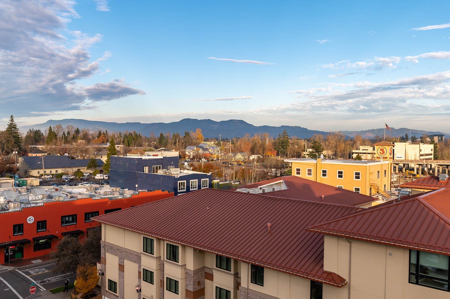 View from Gordon Lofts at the 5th Street Public Market in Eugene, Oregon. The scene features colorful rooftops, including the market's signature red and orange hues, surrounded by a mix of modern and historic architecture. The Cascade foothills provide a stunning backdrop under a clear blue sky with soft clouds.