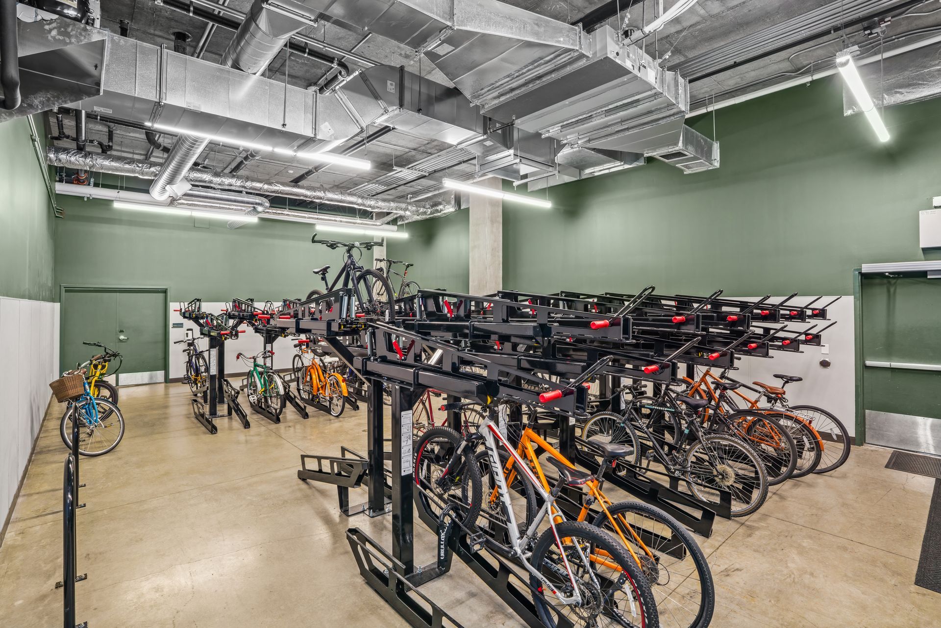 Modern indoor bike storage room at Gordon Lofts in Eugene, Oregon, featuring two-tiered black metal racks with red handles holding a variety of bicycles. The room has polished concrete floors, green and white walls, and exposed ductwork on the ceiling, with bright overhead lighting.