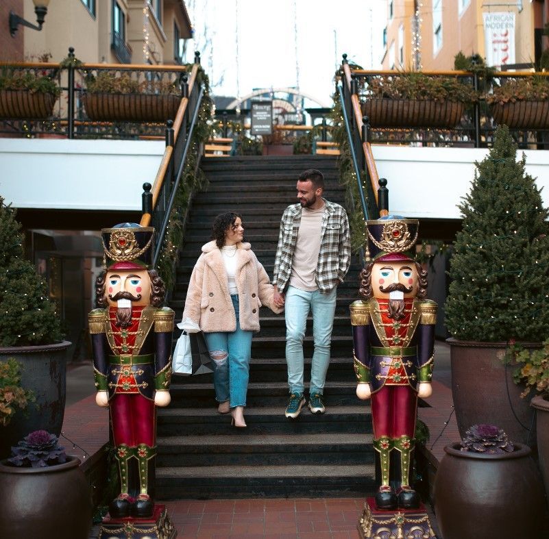 A couple holding hands descends a festive staircase adorned with holiday decorations, including Nutcracker statues and garlands, at 5th Street Public Market, creating a warm, seasonal ambiance.