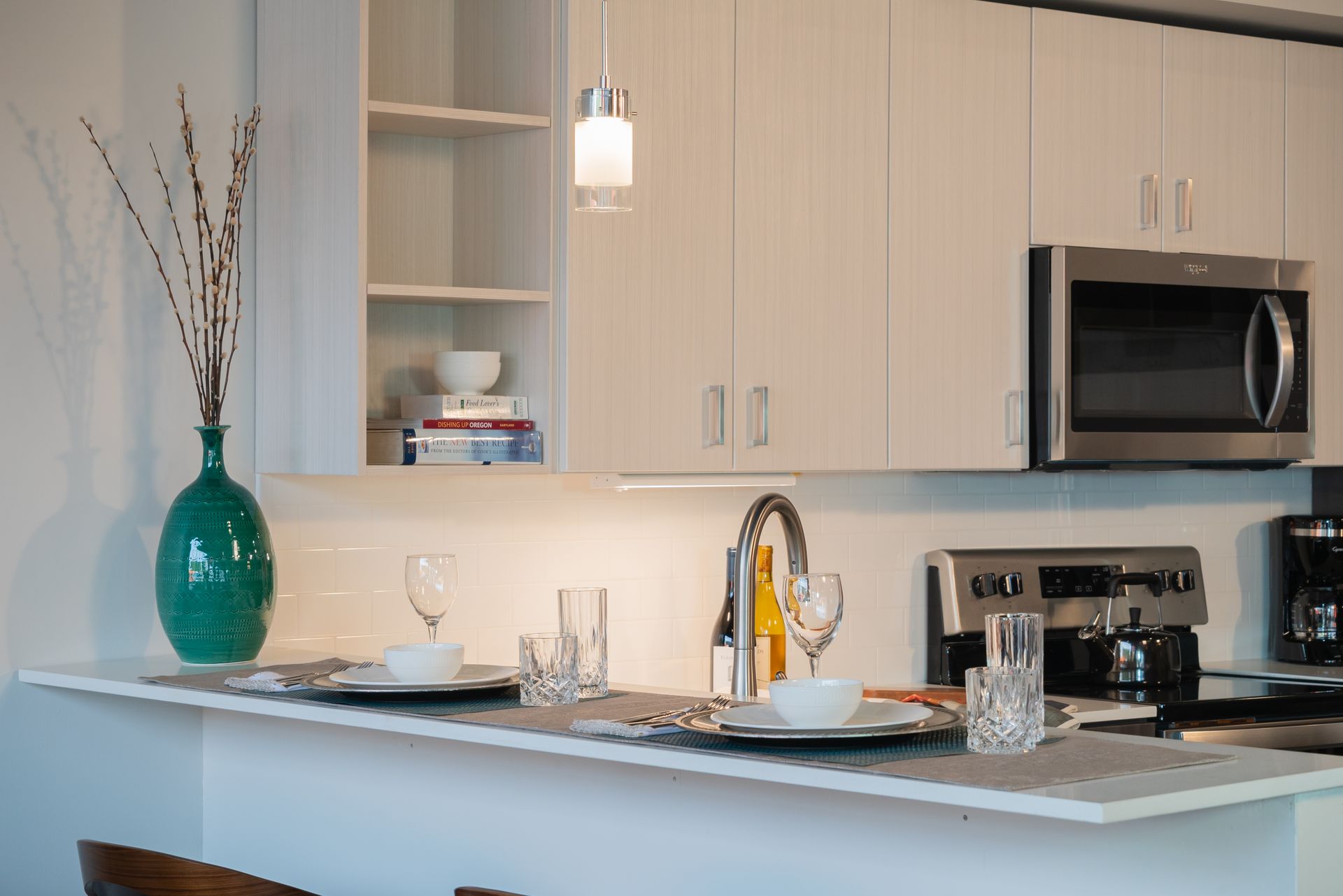 A stylish kitchen setup in the Gordon Lofts showroom. The space features modern cabinetry, stainless steel appliances, and a sleek countertop with a dining setup for two, including plates, glasses, and utensils. A teal vase with decorative branches and neatly stacked books add a touch of elegance and warmth to the design.