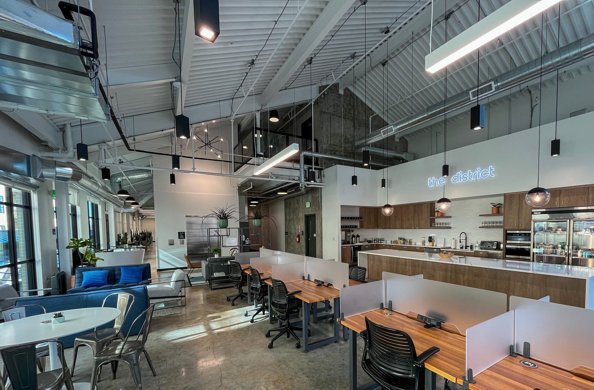 Bright, open-concept coworking space inside The District, directly connected to Gordon Lofts in Eugene, OR. The space features shared desks with frosted dividers, a modern kitchen with bar seating, cozy lounge areas, and floor-to-ceiling windows. Industrial ceilings and pendant lights create a stylish, functional atmosphere ideal for focused work—without ever stepping outside on rainy days.