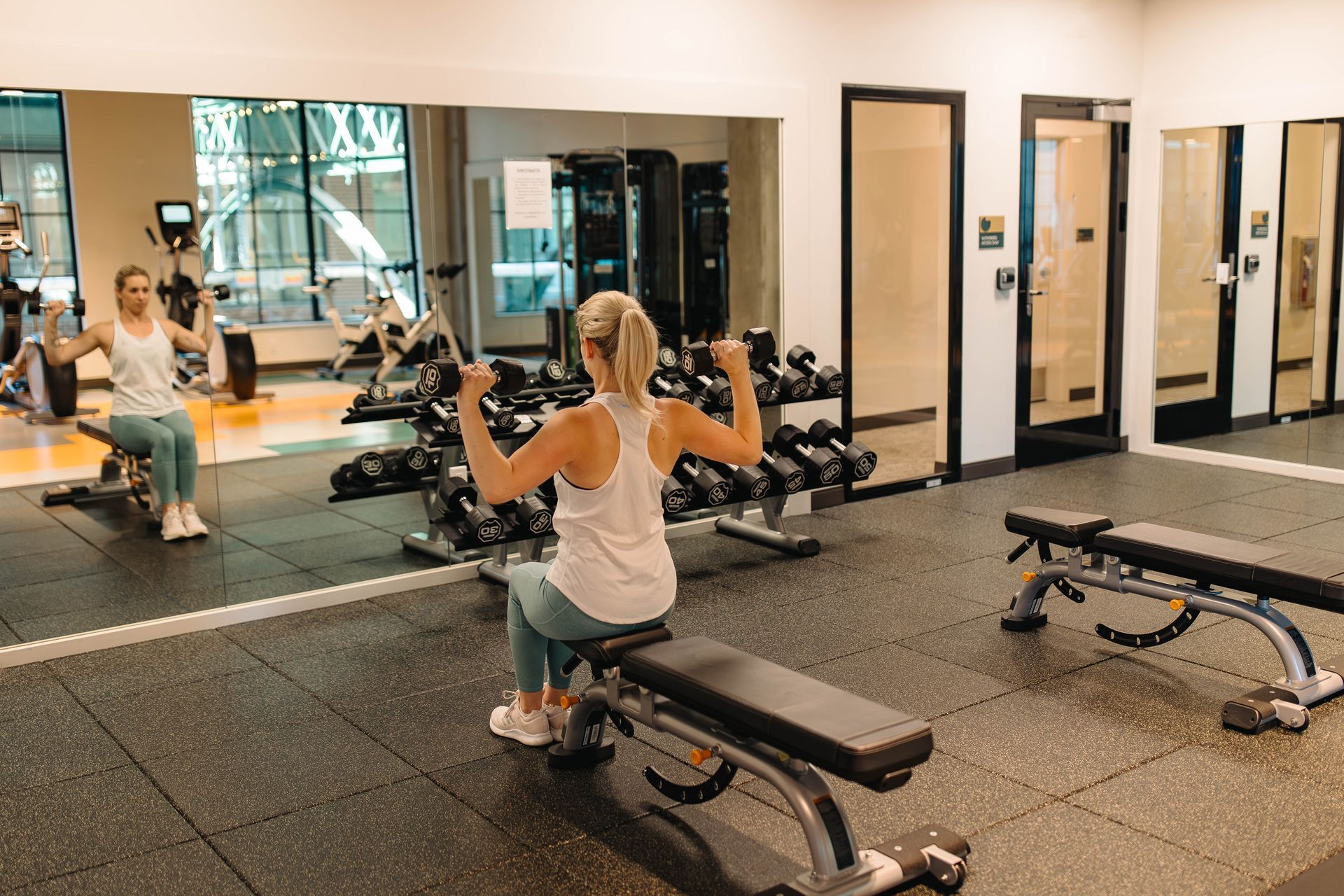 A woman working out in the 24-hour fitness center at Gordon Lofts. She is seated on a bench lifting dumbbells in front of a large mirror, surrounded by modern equipment and a rack of weights. The space is well-lit and inviting, with a clean and organized layout.