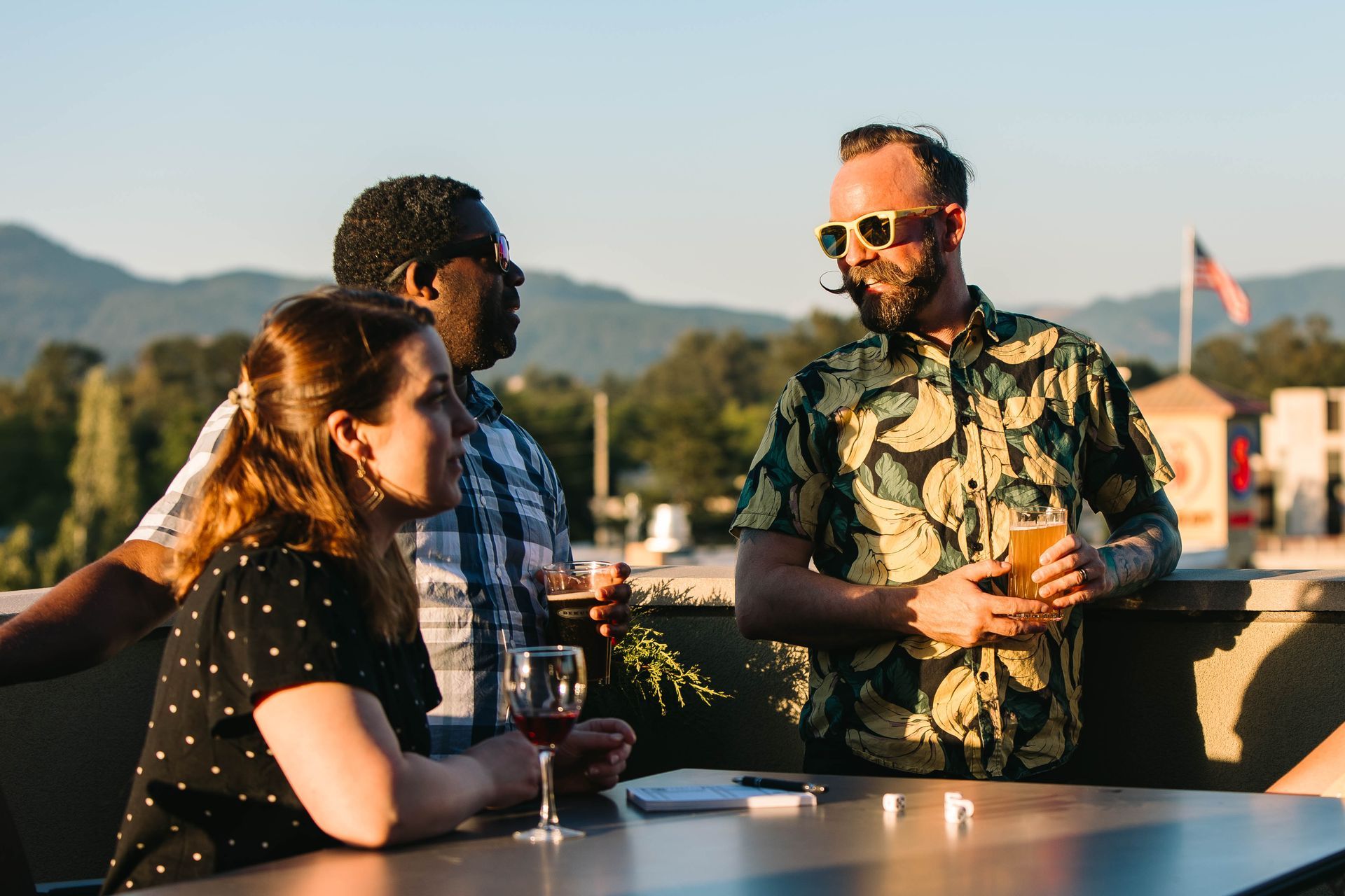 A group of friends enjoying a sunny evening on the Gordon Lofts rooftop lounge. They are gathered around a table with drinks, laughing and chatting, with scenic mountain views and the American flag visible in the background. The relaxed atmosphere highlights the lounge’s appeal for socializing and unwinding.