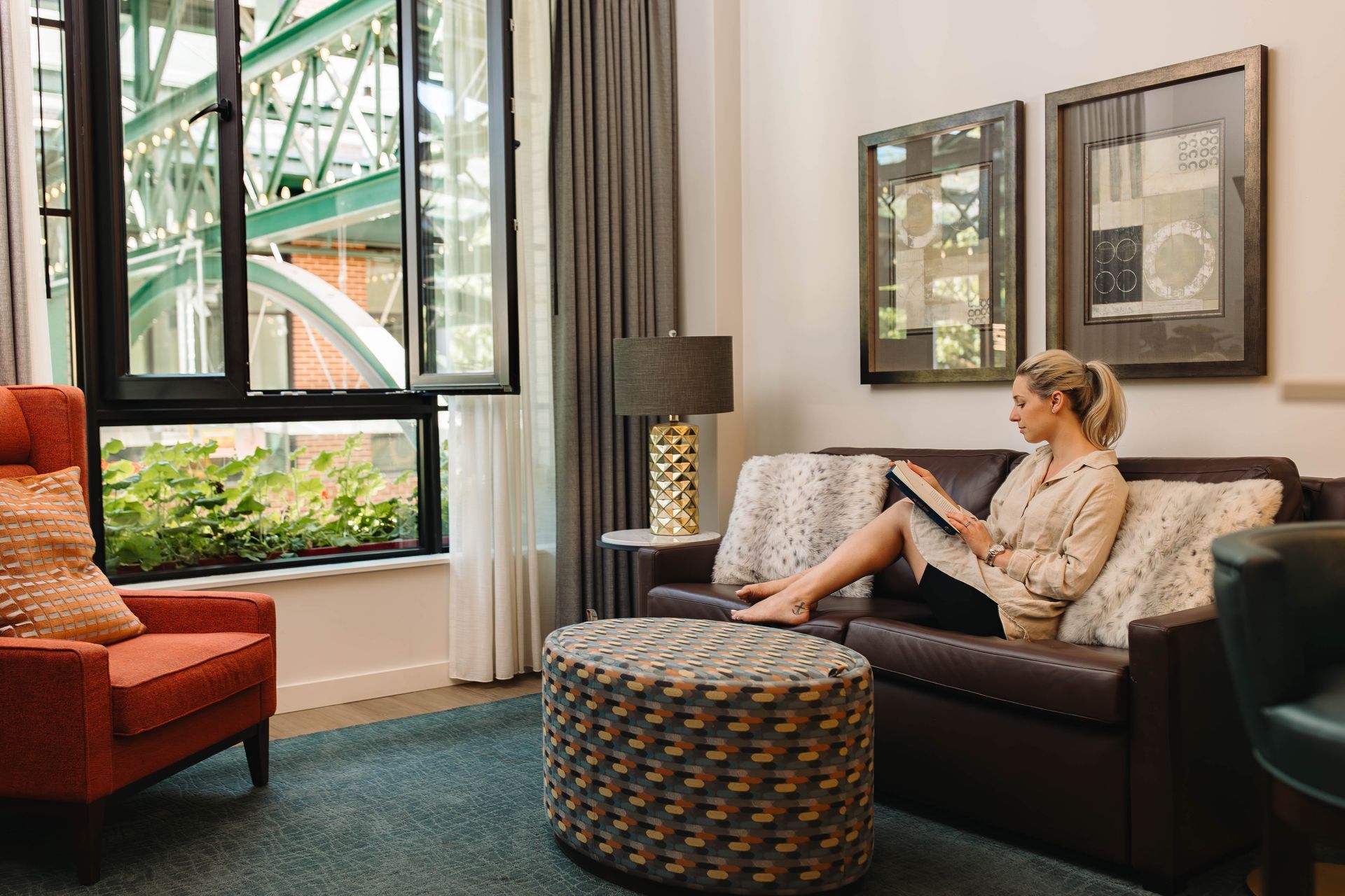 A woman sits comfortably on a dark brown leather sofa with fluffy white pillows, reading a book in a modern, well-lit unit at Gordon Lofts. The space features large windows overlooking the iconic green metal framework of 5th Street Public Market, a patterned ottoman, and an orange accent chair. A geometric gold lamp and abstract wall art add stylish touches to the cozy environment.