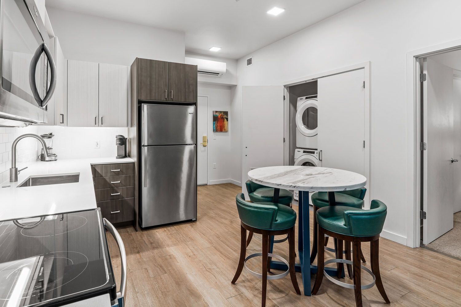 Contemporary kitchen and dining area at Gordon Lofts in Eugene, OR, showcasing stainless steel appliances, white quartz countertops, and wood-styled flooring. A round marble-like topped table with four green upholstered chairs provides a stylish dining space. A stacked washer and dryer are neatly tucked into a closet, highlighting the unit’s convenience and functionality.