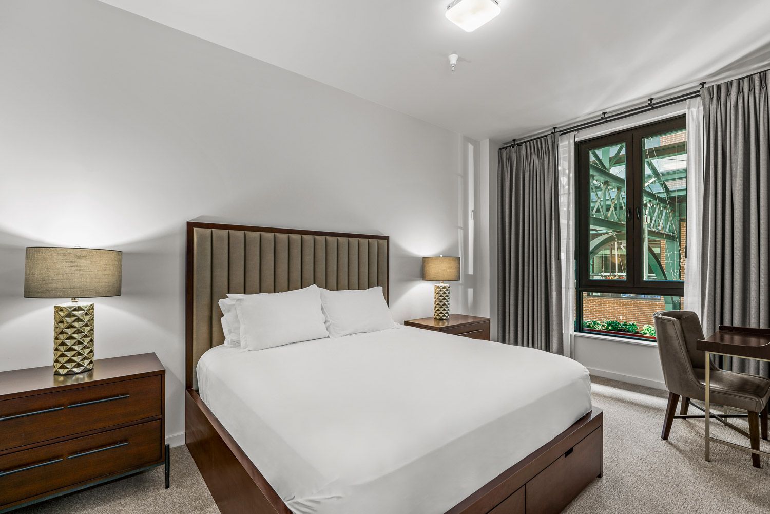 Elegant bedroom at Gordon Lofts in Eugene, OR, featuring a tufted headboard, crisp white bedding, and matching wood nightstands with textured gold lamps. A large window with gray curtains provides a view of the Market District’s distinctive green steel beams. The room also includes a sleek wood desk and chair, blending comfort and sophistication.