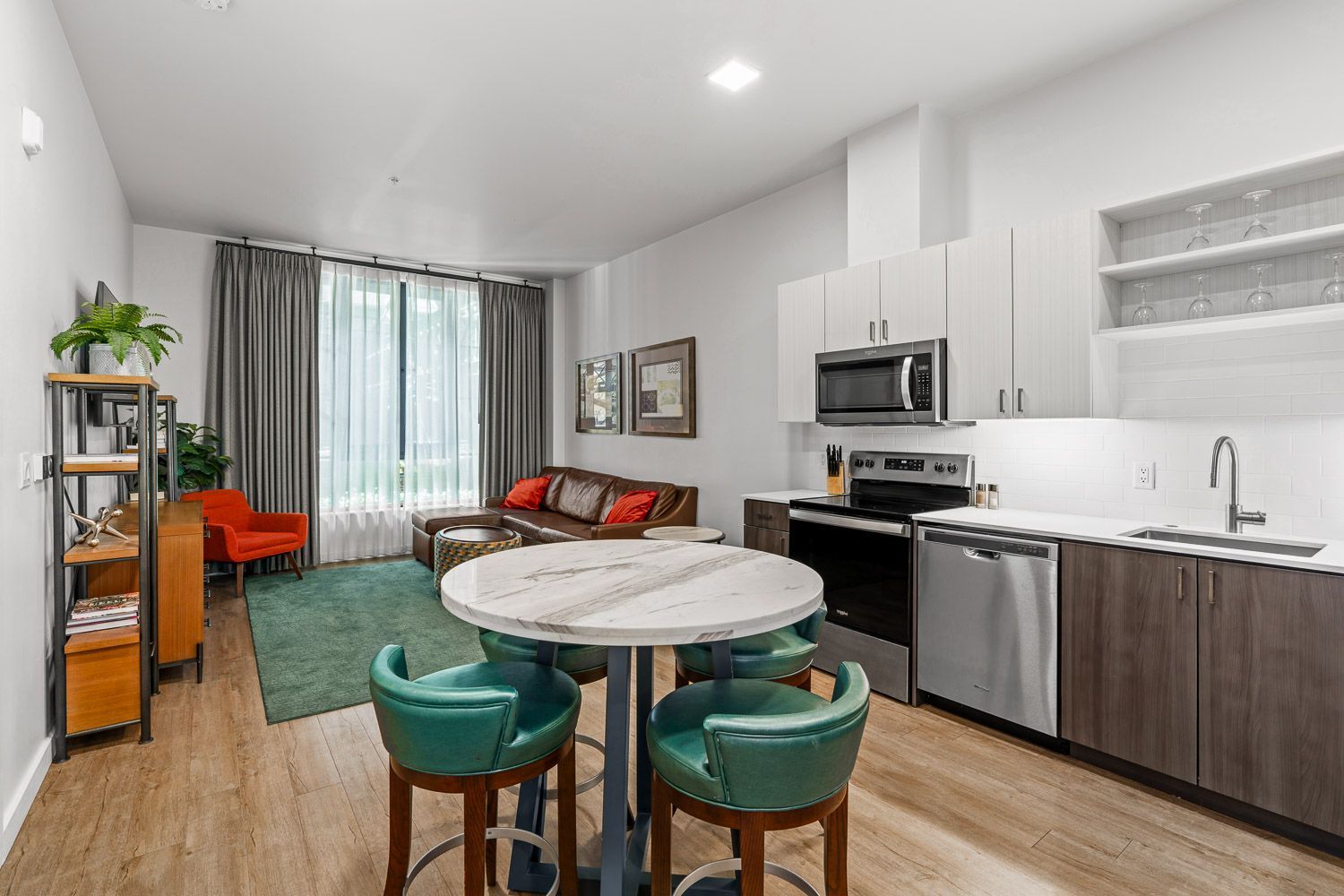 Open-concept kitchen and living space at Gordon Lofts in Eugene, OR, featuring a modern design with a marble-top dining table and four green upholstered chairs. The kitchen includes stainless steel appliances, two-tone cabinetry, and open shelving. The living area offers a brown leather sectional, orange accent chair, green area rug, and large window with floor-to-ceiling curtains, creating a vibrant and comfortable atmosphere.