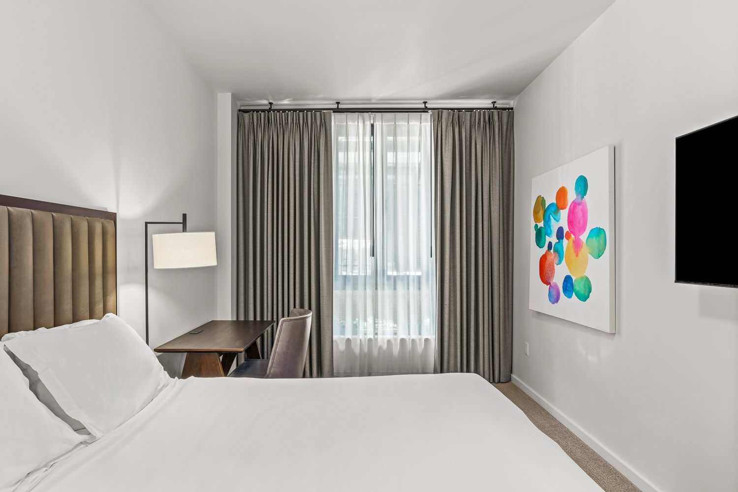 Modern bedroom at Gordon Lofts in Eugene, OR, featuring a plush upholstered headboard, crisp white bedding, and a sleek desk with a reading lamp. The room includes a flat-screen TV, contemporary colorful wall art, and floor-to-ceiling curtains that frame a large window, creating a serene and stylish atmosphere.