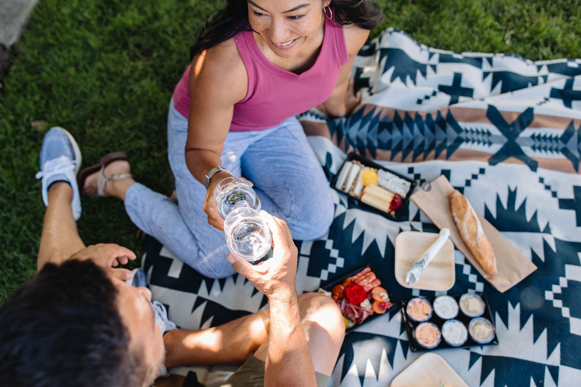 A couple enjoys a picnic on a patterned blanket spread over green grass at the 5th Street Public Market. They are toasting with glasses of water, surrounded by a spread of cheeses, dips, charcuterie, and a baguette. The woman is smiling and wearing a pink tank top and striped pants, while the man is seated beside her in casual summer clothes.