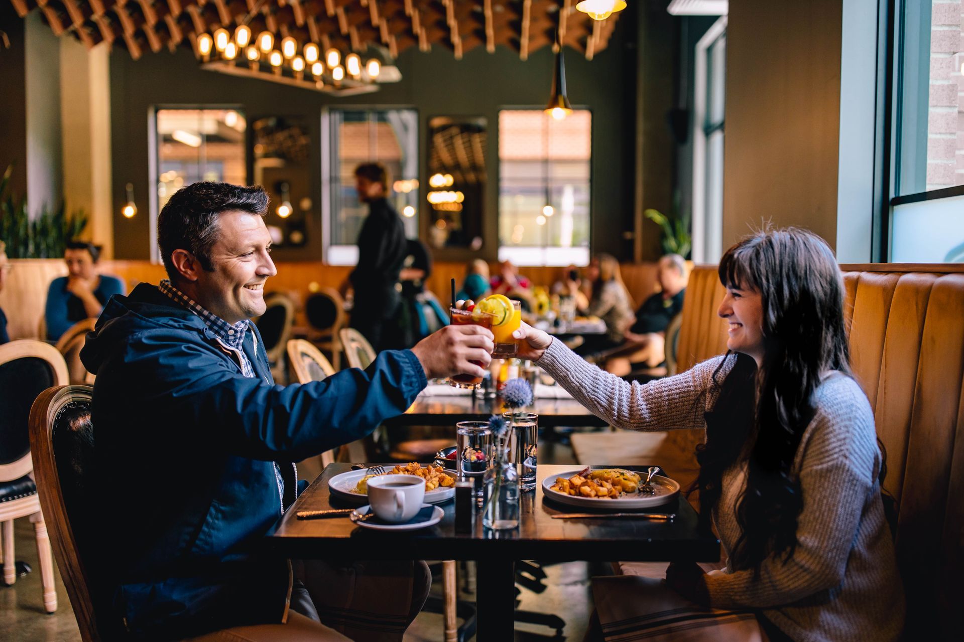 A cheerful moment at the Gordon Tavern inside The Gordon Hotel in Eugene, Oregon. A man and woman share a toast with vibrant cocktails at a cozy table. The inviting ambiance features warm lighting, stylish decor, and a lively dining atmosphere with other patrons in the background.