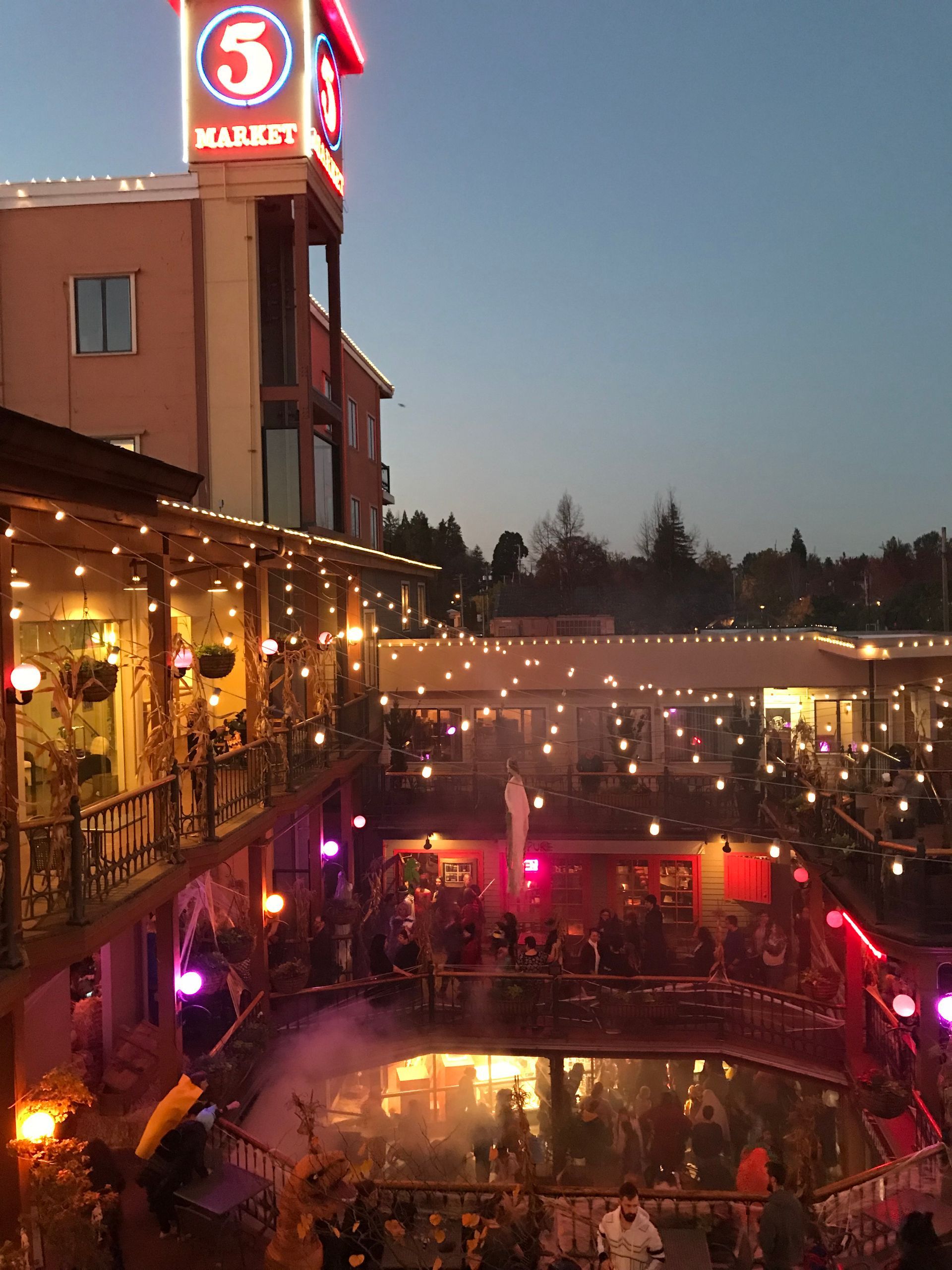 A festive evening scene at the Haunted Market event in the Market Core of Eugene’s 5th Street Public Market. The courtyard is aglow with string lights, purple and orange lanterns, and Halloween-themed decor, including fog effects and a ghostly figure suspended in the center. Families and costumed guests fill the multi-level space, creating a lively and whimsical atmosphere beneath the iconic illuminated “5” tower.