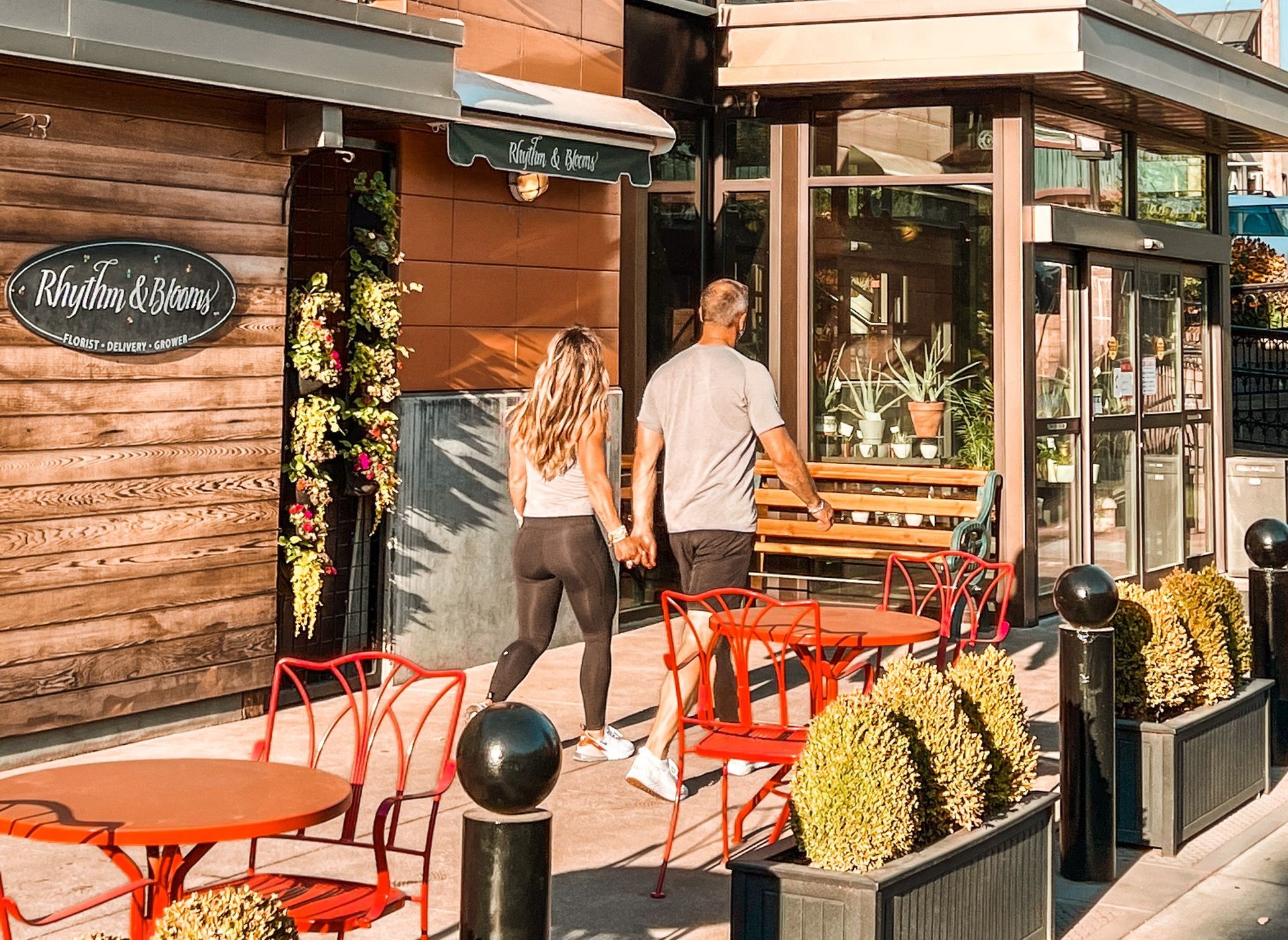 A couple holds hands as they walk toward the entrance of Rhythm and Blooms, a florist shop located in the 5th Street Public Market in Eugene, Oregon. The storefront features warm wood paneling, a green awning, and large windows displaying potted plants. Bright red patio chairs and tables line the sidewalk, with manicured shrubs and blooming flowers adding to the inviting outdoor setting on a sunny day.