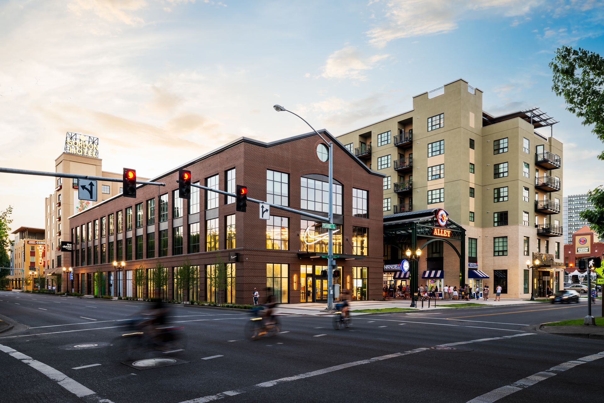 Market Alley and the Gordon Lofts in downtown Eugene, Oregon. The Nike Store is prominently featured with its sleek, modern design. The area showcases the vibrant and bustling atmosphere of the 5th Street Public Market, with people enjoying nearby shops and restaurants under a beautiful evening sky.