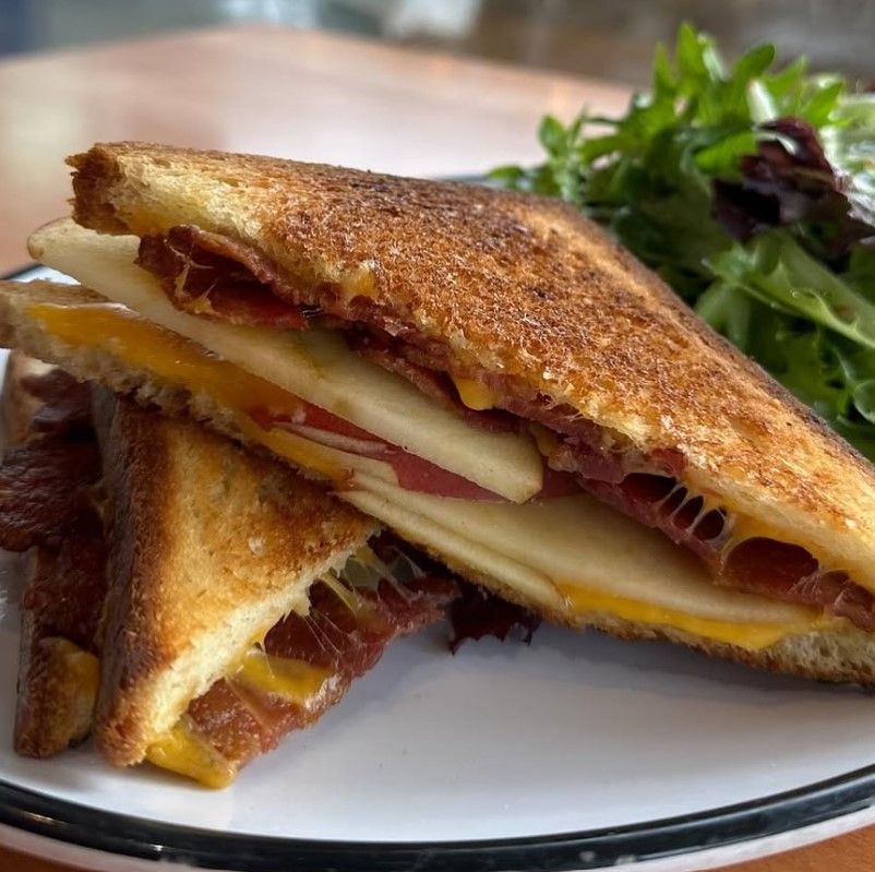 A gourmet grilled cheese sandwich from Provisions at 5th Street Public Market, featuring crispy golden-brown toasted bread filled with melted cheddar cheese, thinly sliced apples, and crispy bacon. The sandwich is cut in half, showcasing the layers of sweet and savory flavors. It is served with a side of fresh mixed greens for a balanced and delicious meal.