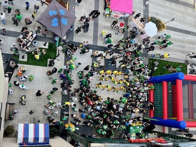 A bird's-eye view of a lively 'Rally in the Alley' event at 5th Street Public Market, featuring a crowd of people in green and yellow attire, outdoor tents, a bounce house, and vendor booths.