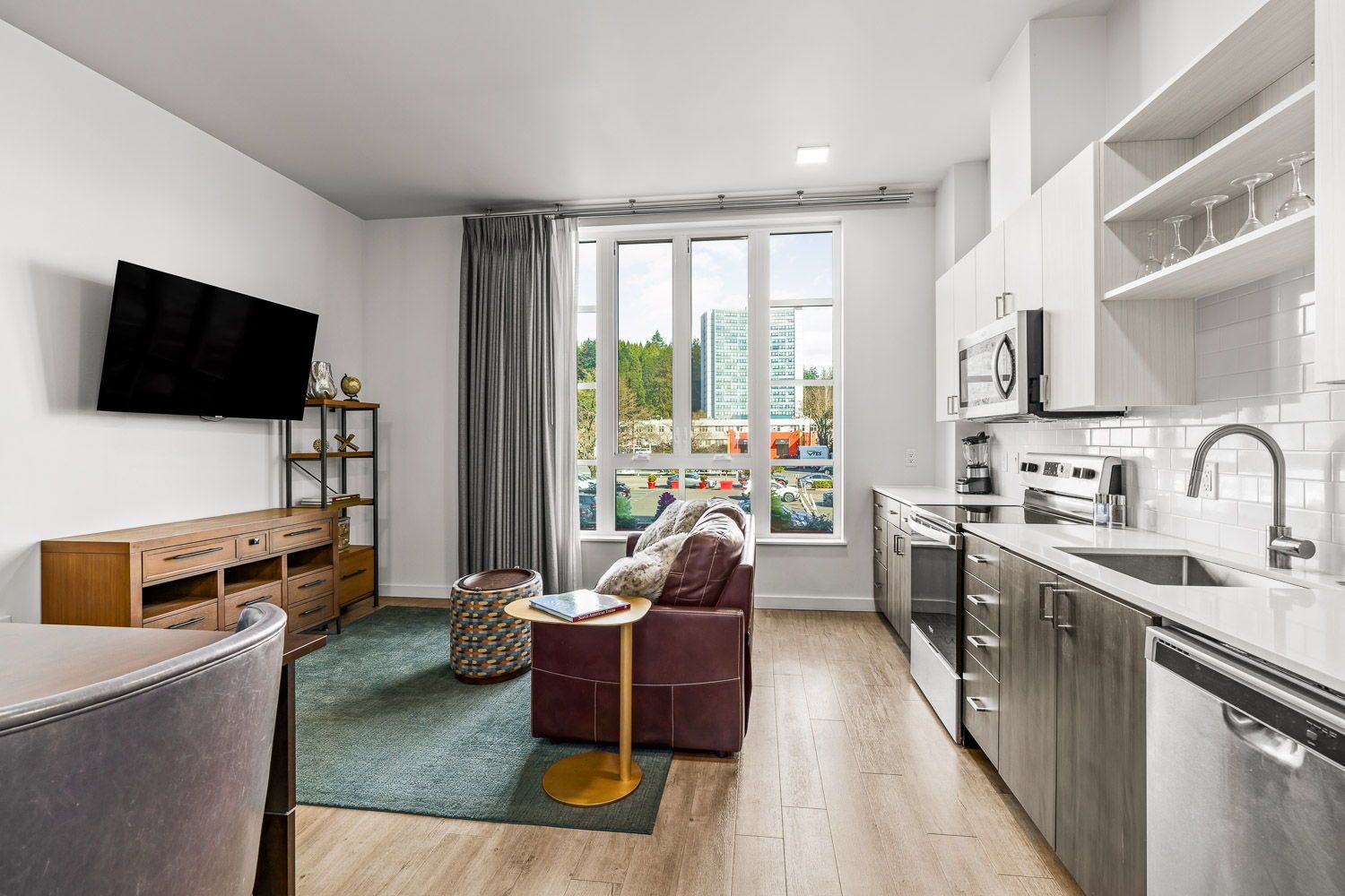 Stylish furnished apartment at Gordon Lofts in Eugene, OR, featuring a modern kitchen with stainless steel appliances, open shelving, and subway tile backsplash. The cozy living area includes a mounted flat-screen TV, leather armchair, small round table, and large windows offering natural light and a view of the 5th Street Public Market area.