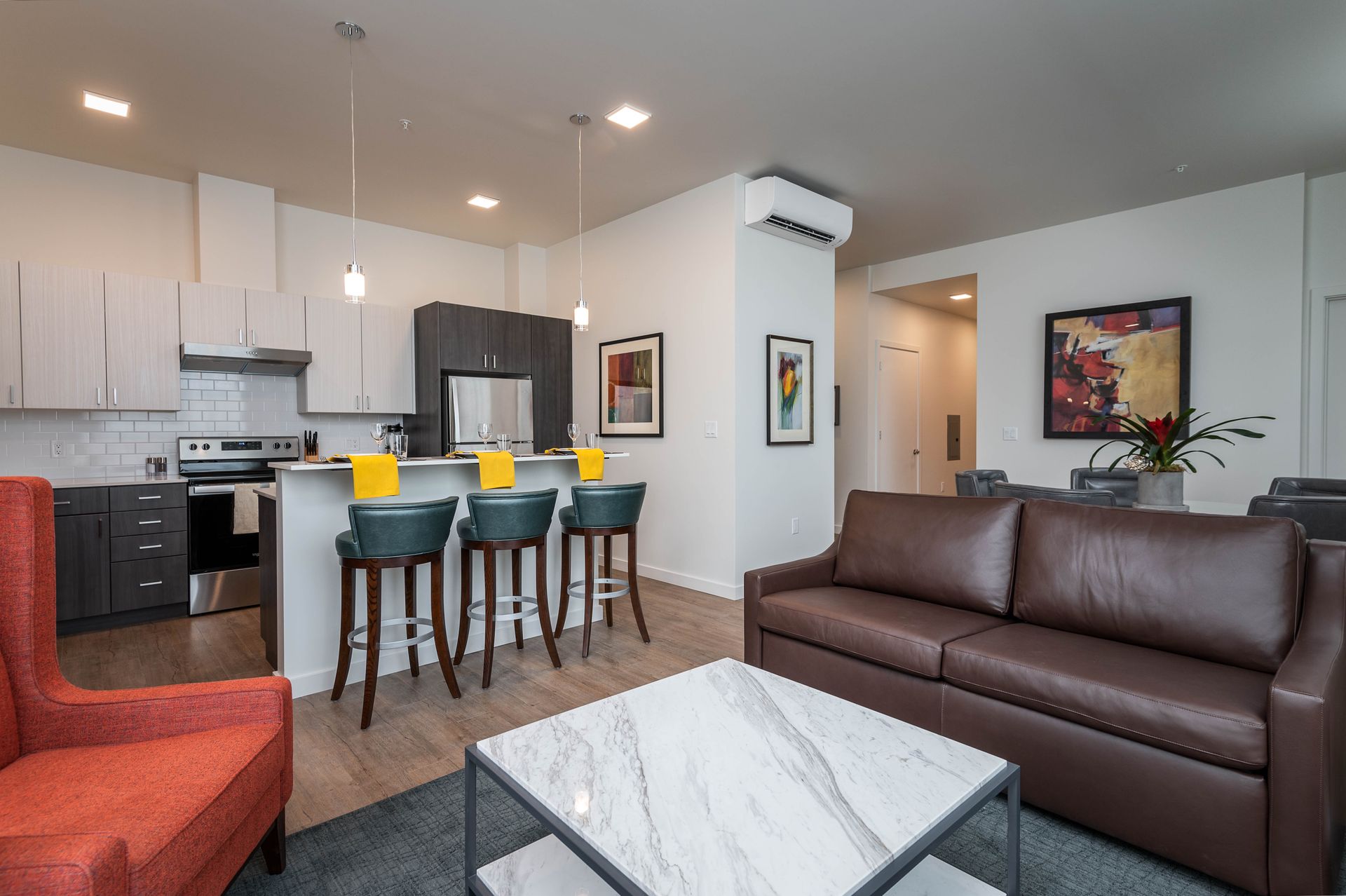 A modern and fully furnished living space in Unit 201 at the Gordon Lofts. The room features a sleek kitchen with stainless steel appliances, a marble coffee table, vibrant red and brown seating, and contemporary artwork, offering a stylish and welcoming atmosphere.