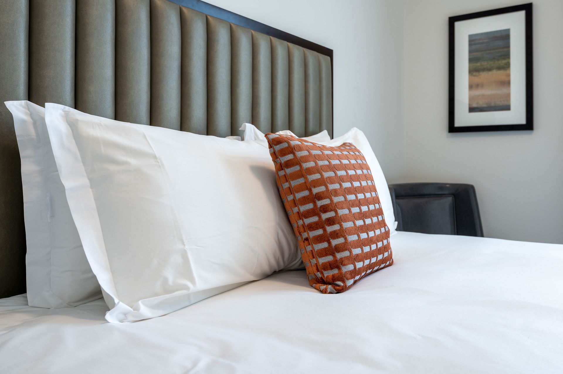 A cozy bedroom setup in furnished unit 201 at Gordon Lofts. The bed features crisp white linens, an accent pillow with an orange and gray pattern, and a modern upholstered headboard. A framed artwork and a sleek gray chair add a touch of sophistication to the minimalist decor.