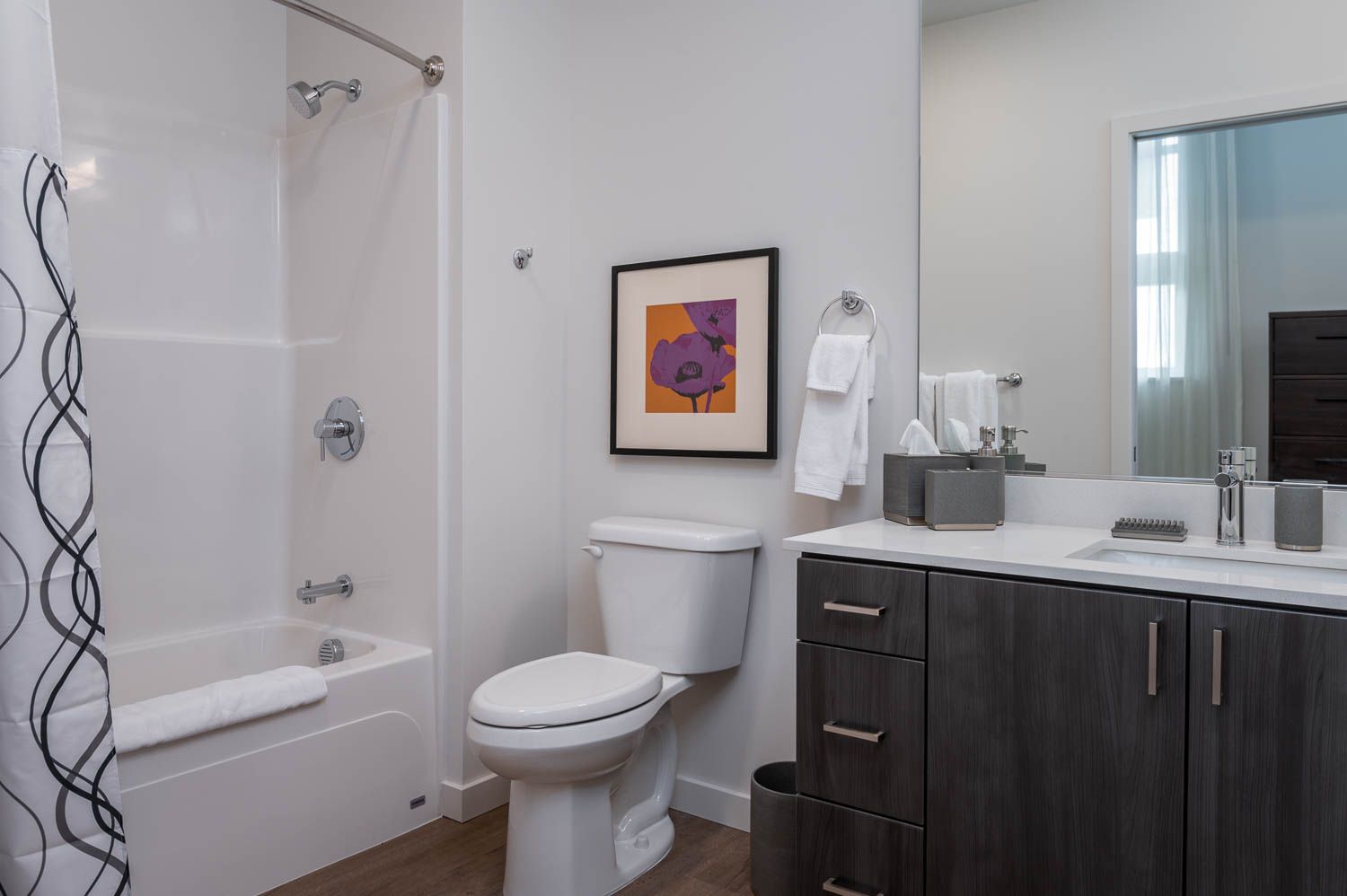 A sleek and modern bathroom in furnished unit 201 at Gordon Lofts. The space features a bathtub with a curved shower rod and a patterned shower curtain, a white toilet, and a vanity with dark wood cabinetry and a quartz countertop. A vibrant framed artwork adds a pop of color, while neatly arranged towels and accessories create a polished, inviting look.