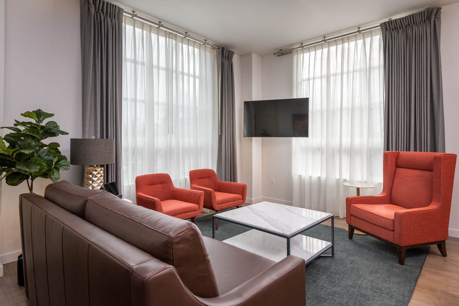 A modern and elegant living space in furnished unit 201 at Gordon Lofts. The room features tall windows with sheer and gray curtains, a sleek brown leather sofa, vibrant orange armchairs, and a matching wingback chair. A marble coffee table and a mounted TV complete the sophisticated look, accented by a stylish floor lamp and a lush green plant.