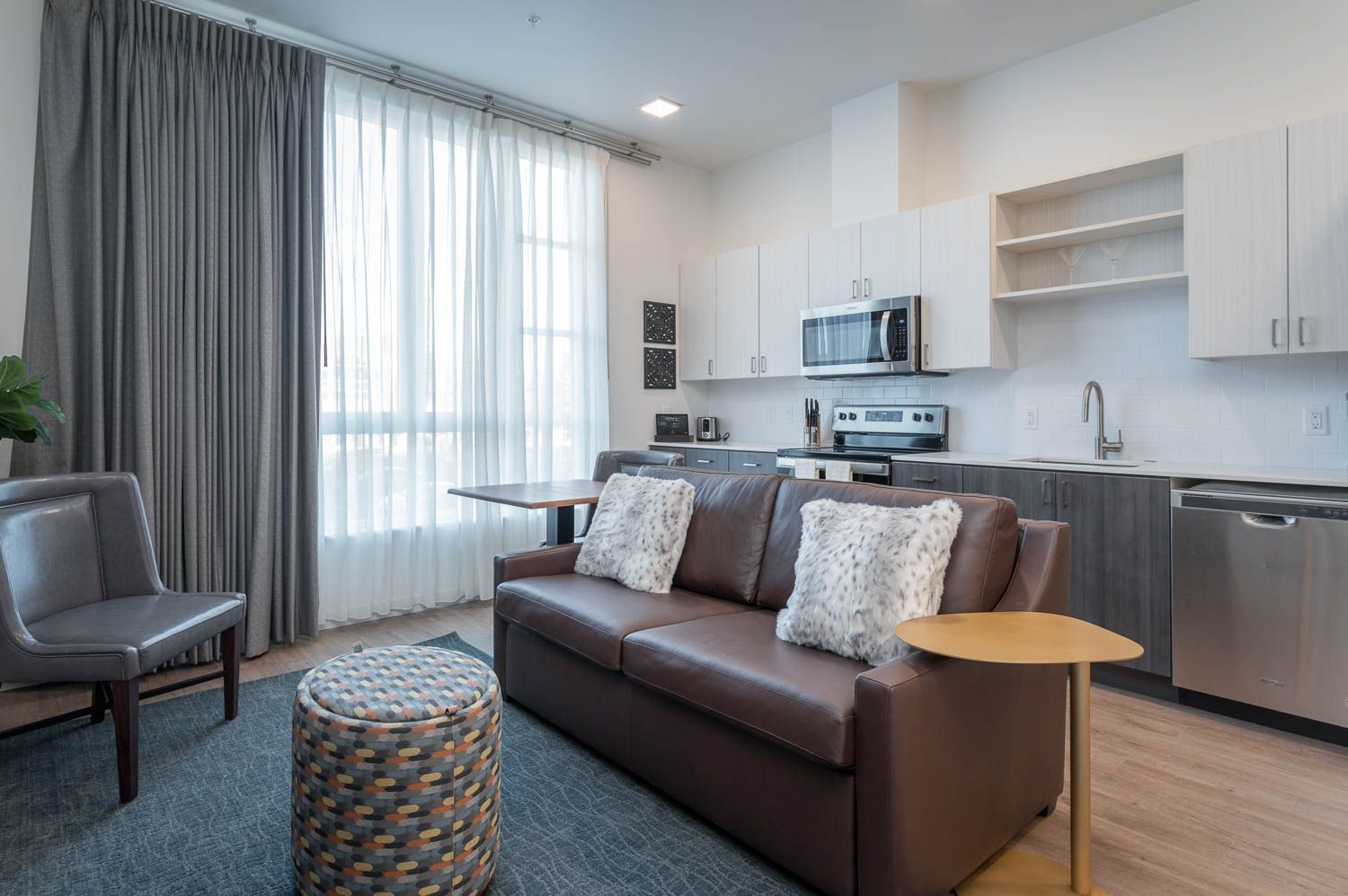 A modern living space in furnished unit 207 at Gordon Lofts. The room features a cozy brown leather sofa with faux fur pillows, a patterned ottoman, and a sleek gray chair. Large windows with sheer and gray curtains bring in natural light. The open kitchen includes white and gray cabinetry, stainless steel appliances, and a clean, minimalist design.