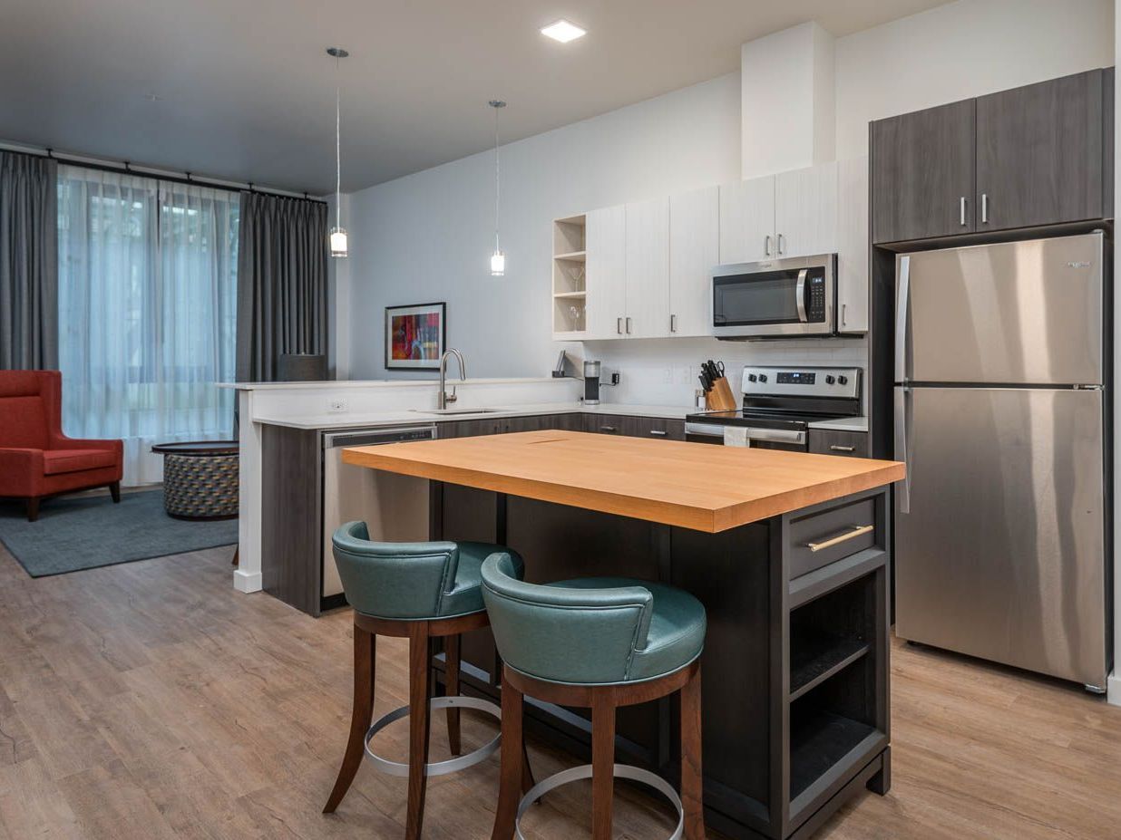 A spacious and modern kitchen in furnished unit 214 at Gordon Lofts. The kitchen features a large island with a wood countertop and teal barstools, stainless steel appliances, and sleek cabinetry in a two-tone design. Pendant lighting adds a stylish touch, and the open layout flows into a cozy living area with a red accent chair and large windows.