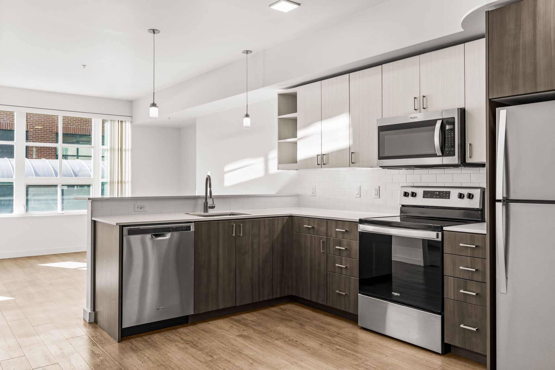 Contemporary corner kitchen at Gordon Lofts in Eugene, OR, featuring stainless steel appliances, modern two-tone cabinetry, white subway tile backsplash, and sleek pendant lighting. The open layout flows into a bright living space with large windows and wood-style flooring, creating a clean and inviting atmosphere.