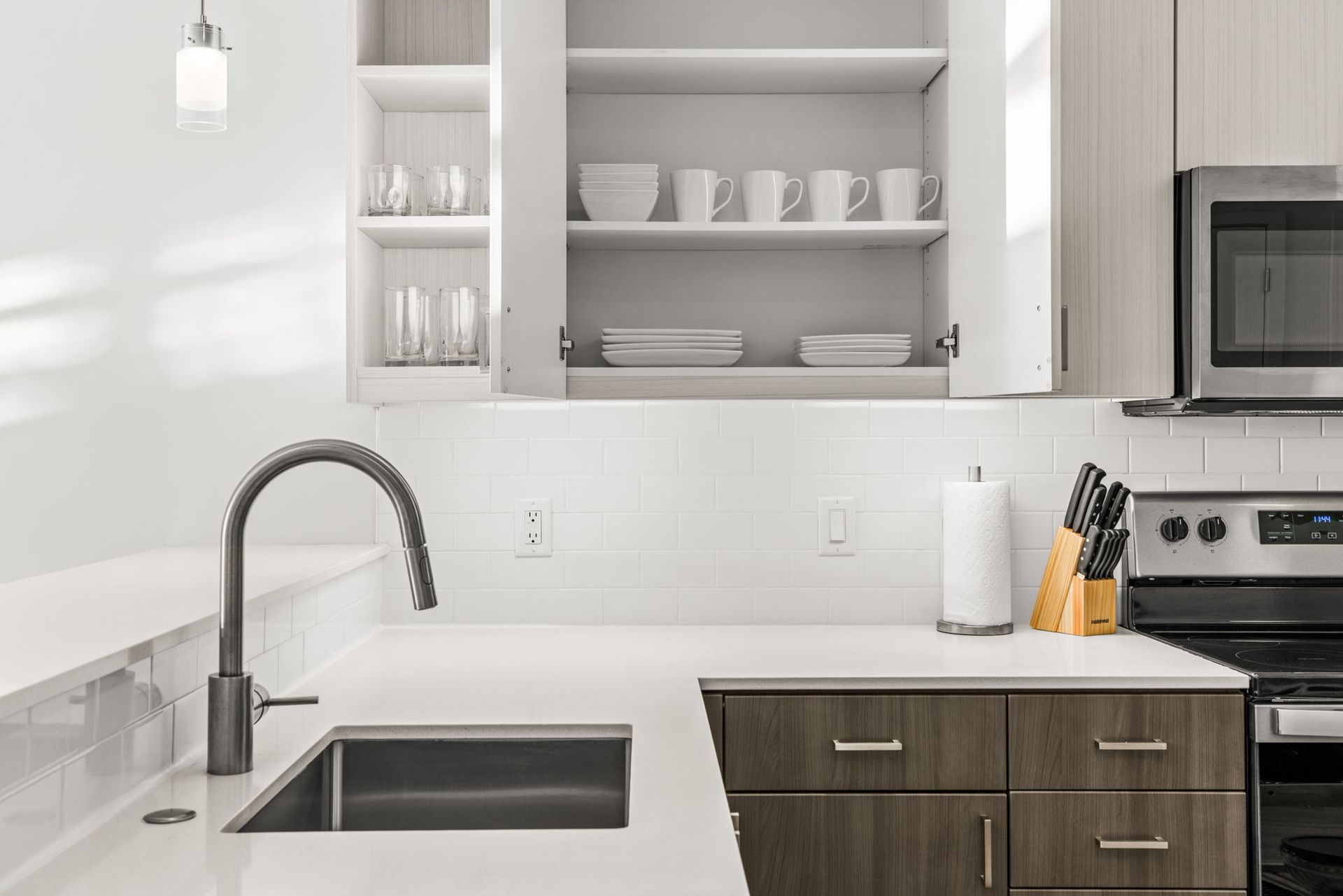 Sleek kitchen at Gordon Lofts in Eugene, OR, featuring a modern design with white quartz countertops, stainless steel appliances, a deep under-mount sink, and minimalist cabinetry. Open upper cabinets display neatly arranged dishes, mugs, and glassware, while a knife block and paper towel holder sit on the counter below. The white subway tile backsplash adds a clean, contemporary touch.