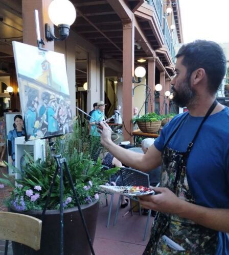 Alejandro painting at Art With Alejandro’s Studio & Gallery at 5th Street Public Market. He is focused on his artwork, holding a palette and brush, with a partially completed painting on an easel. The setting is lively with people and charming outdoor decor.