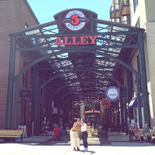 Two people posing and smiling in the center of Market Alley at 5th Street Public Market. The arched entryway with the 'ALLEY' sign and 5th Street Public Market logo is prominently displayed above them. The scene is lively with people dining and walking in the background under the metal-framed structure.