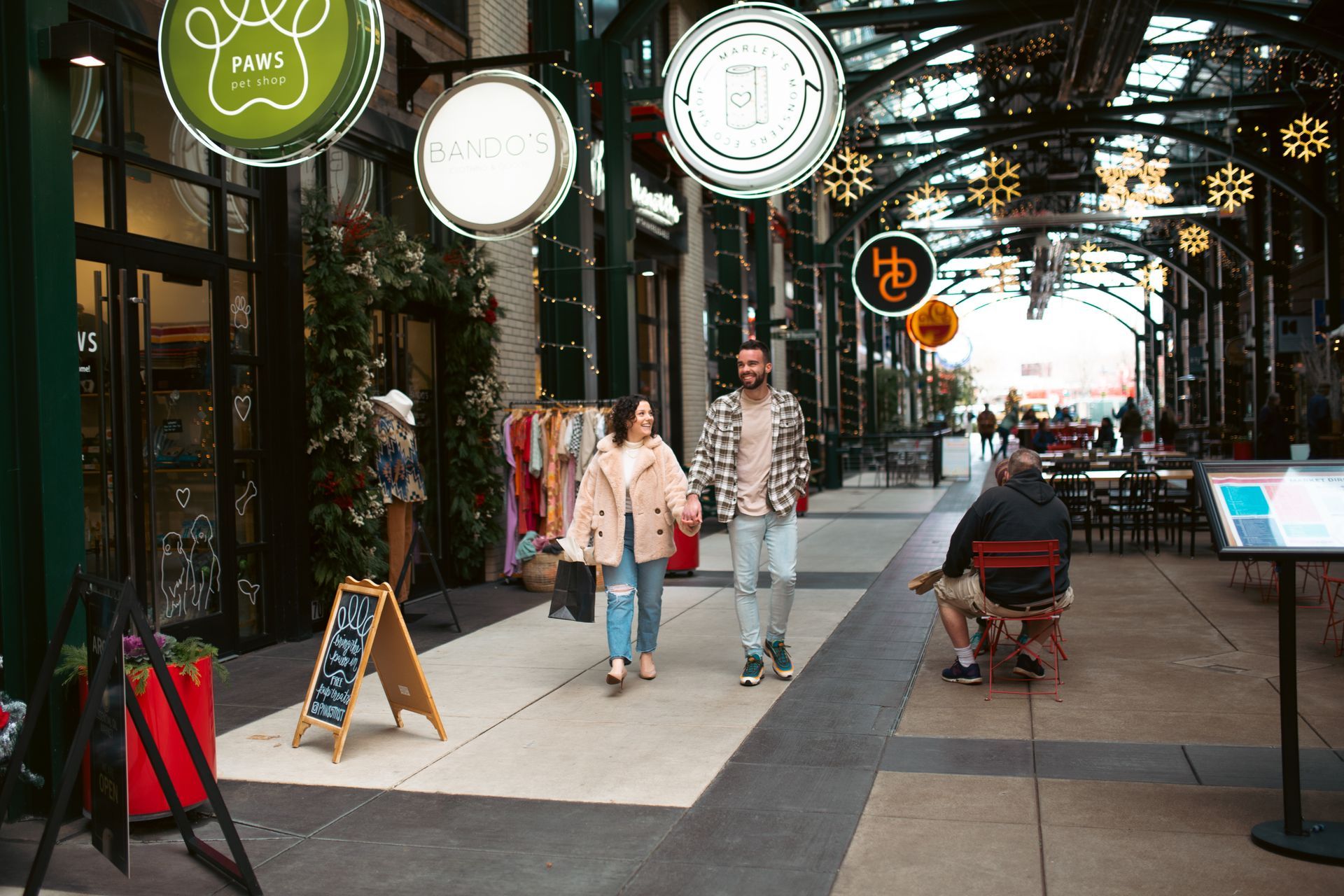 A couple walking hand in hand through Market Alley at 5th Street Public Market. The alley is adorned with festive decorations, including hanging snowflake lights and illuminated shop signs. Stores such as 'Marley's Monsters' and 'Bando’s' are visible along the path, with displays of clothing and accessories outside. The atmosphere is lively, with other visitors sitting and walking in the background, surrounded by decorated planters and a charming marketplace vibe.